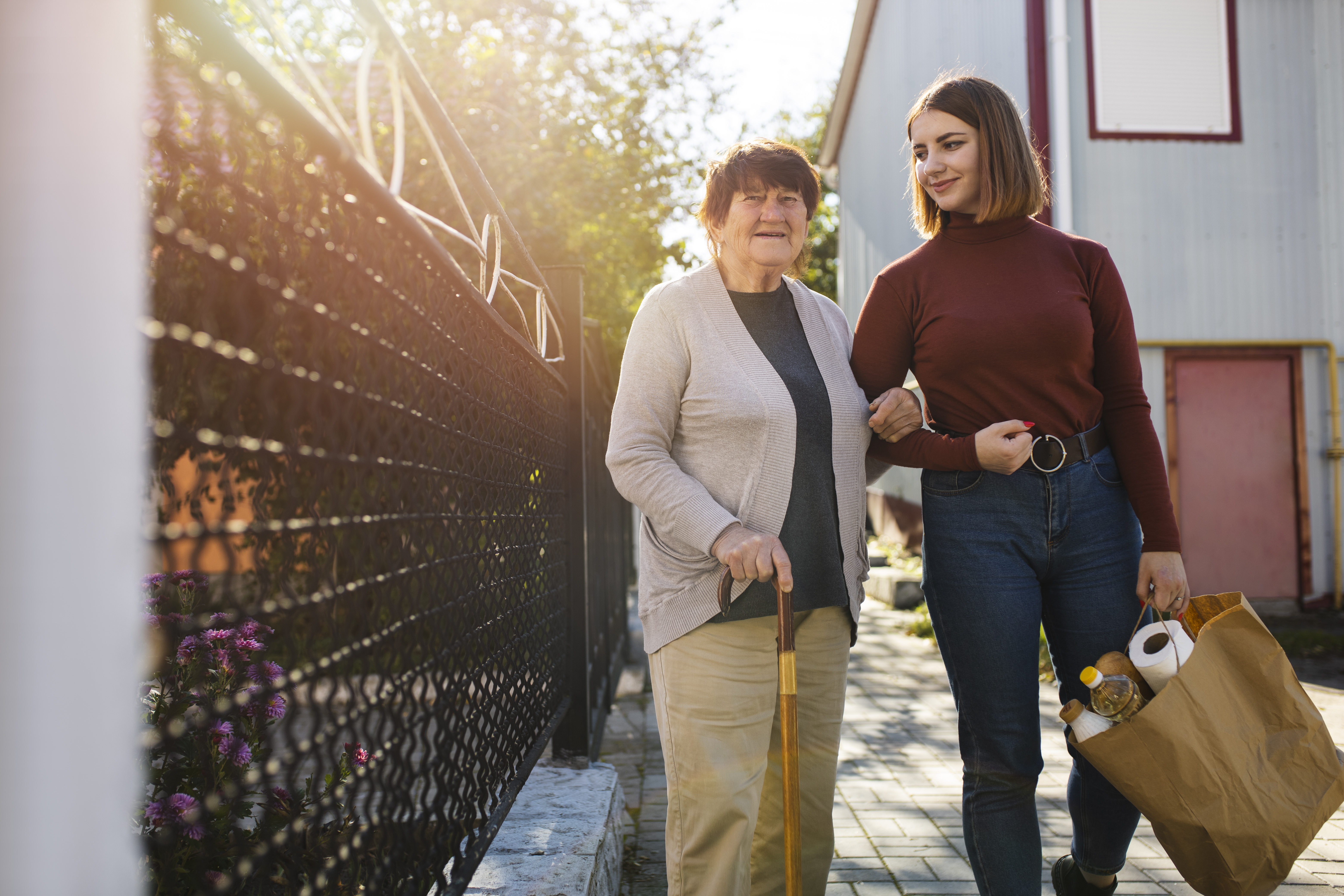 Photo of an older woman with a walking stick walking arm-in-arm along a pathway by a house. The younger woman is carrying a shopping bag.