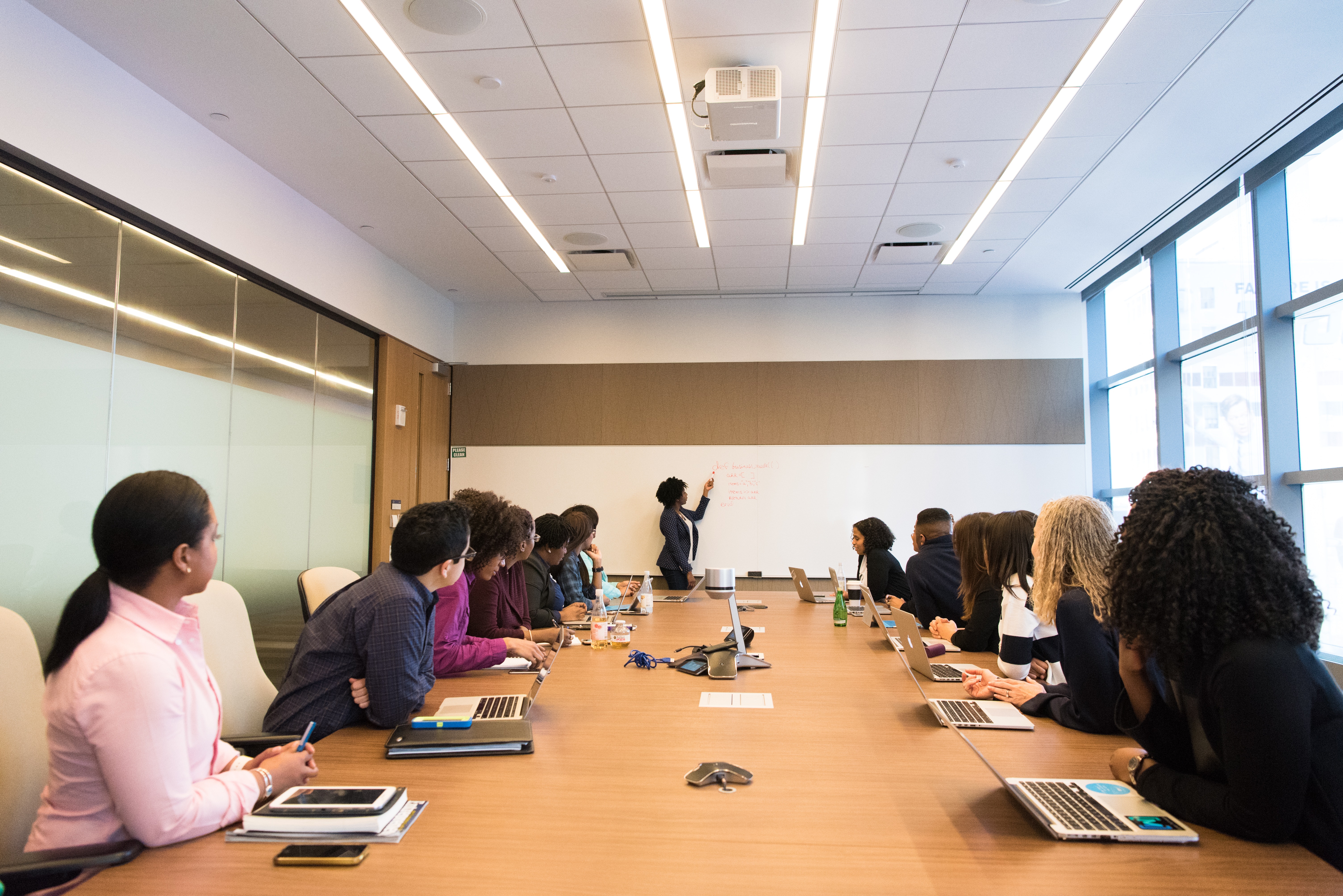 Photograph of several people seated around a long table, each with a laptop in front of them. The photo is taken from a low angle looking along the table. At the far end a woman is writing on a whiteboard