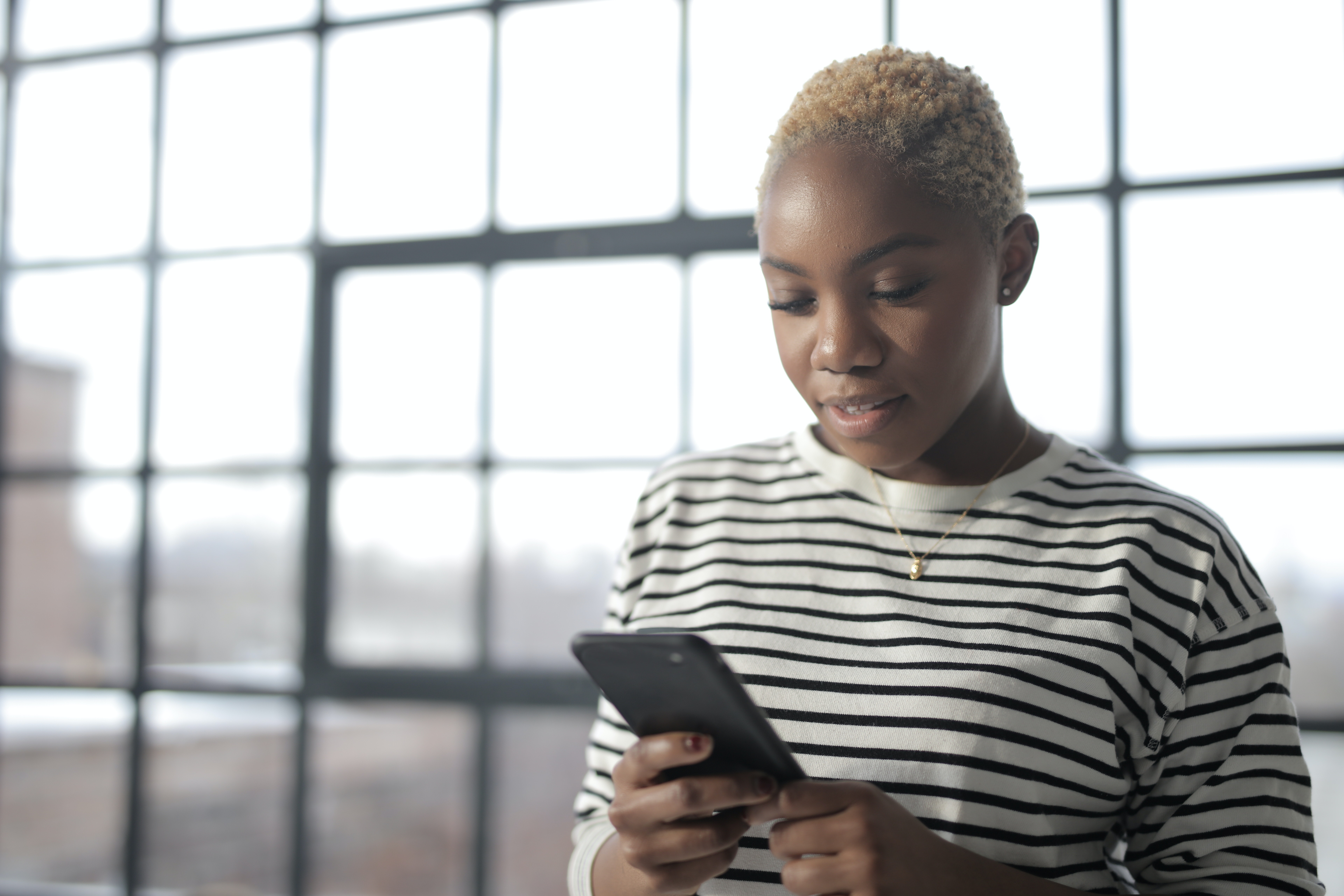Photo of a young black woman with bleached hair looking at her phone. She is standing in front of a large window.