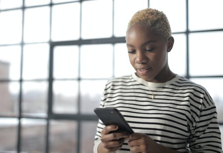 Photo of a young black woman with bleached hair looking at her phone. She is standing in front of a large window.