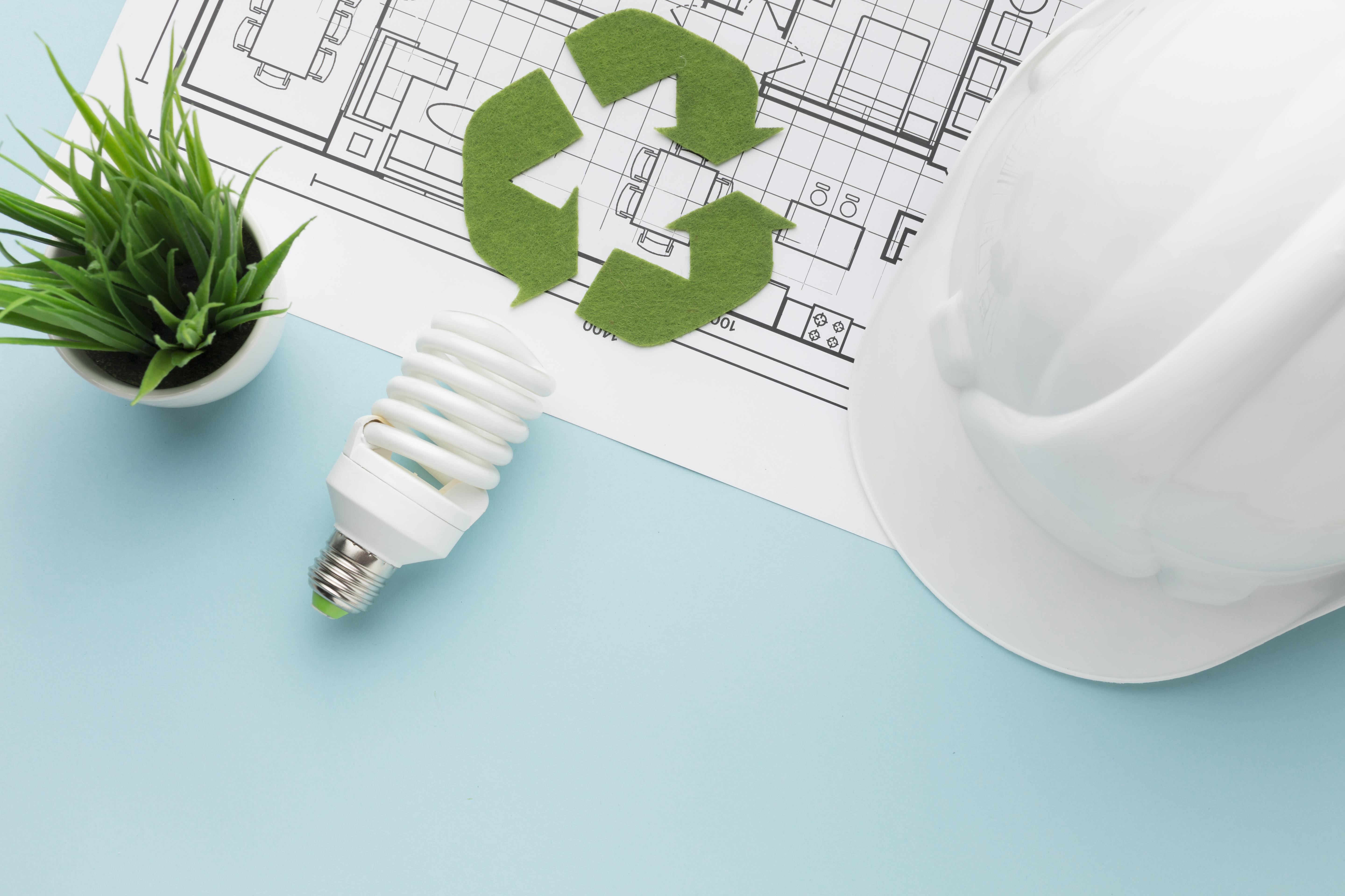 Photo of a white hard hat, a floorplan for a building, a low energy bulb a small plant and a green cut-out recycling symbol lying on a turquoise surface