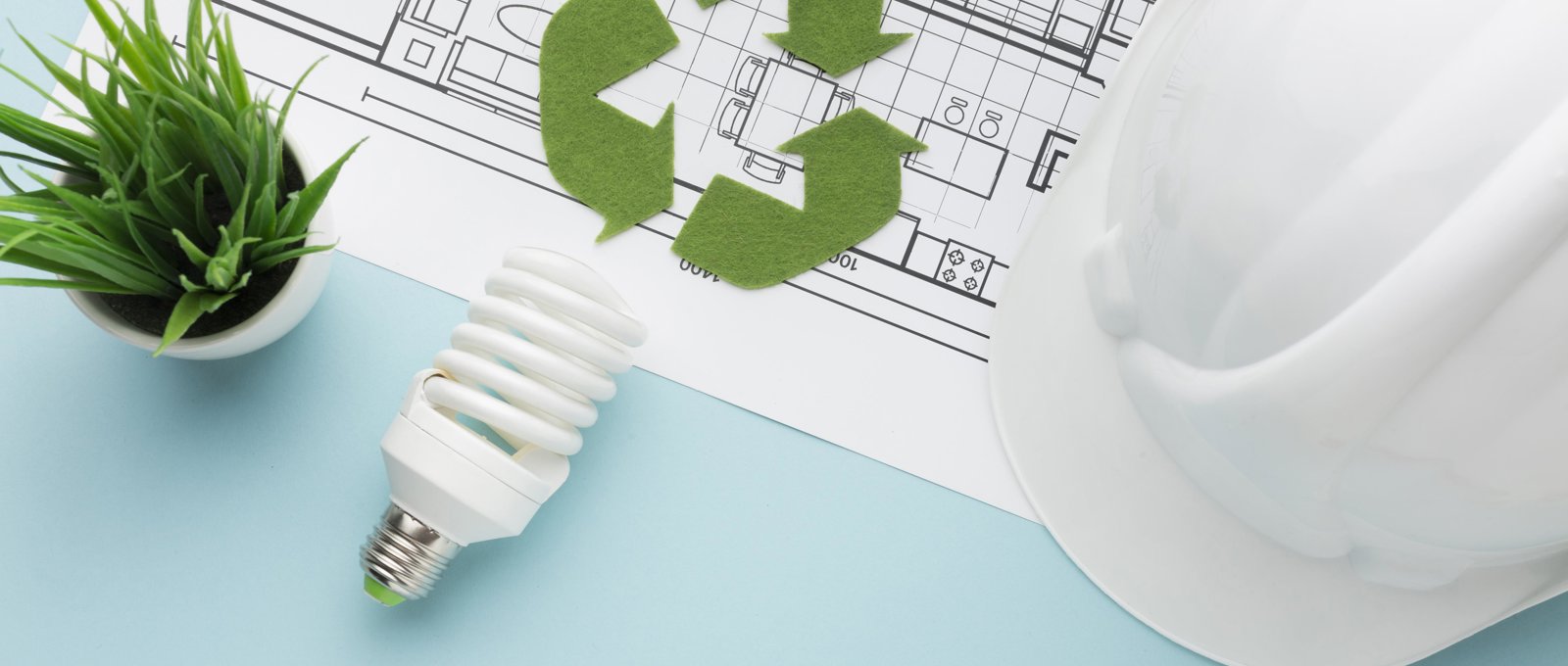 Photo of a white hard hat, a floorplan for a building, a low energy bulb a small plant and a green cut-out recycling symbol lying on a turquoise surface