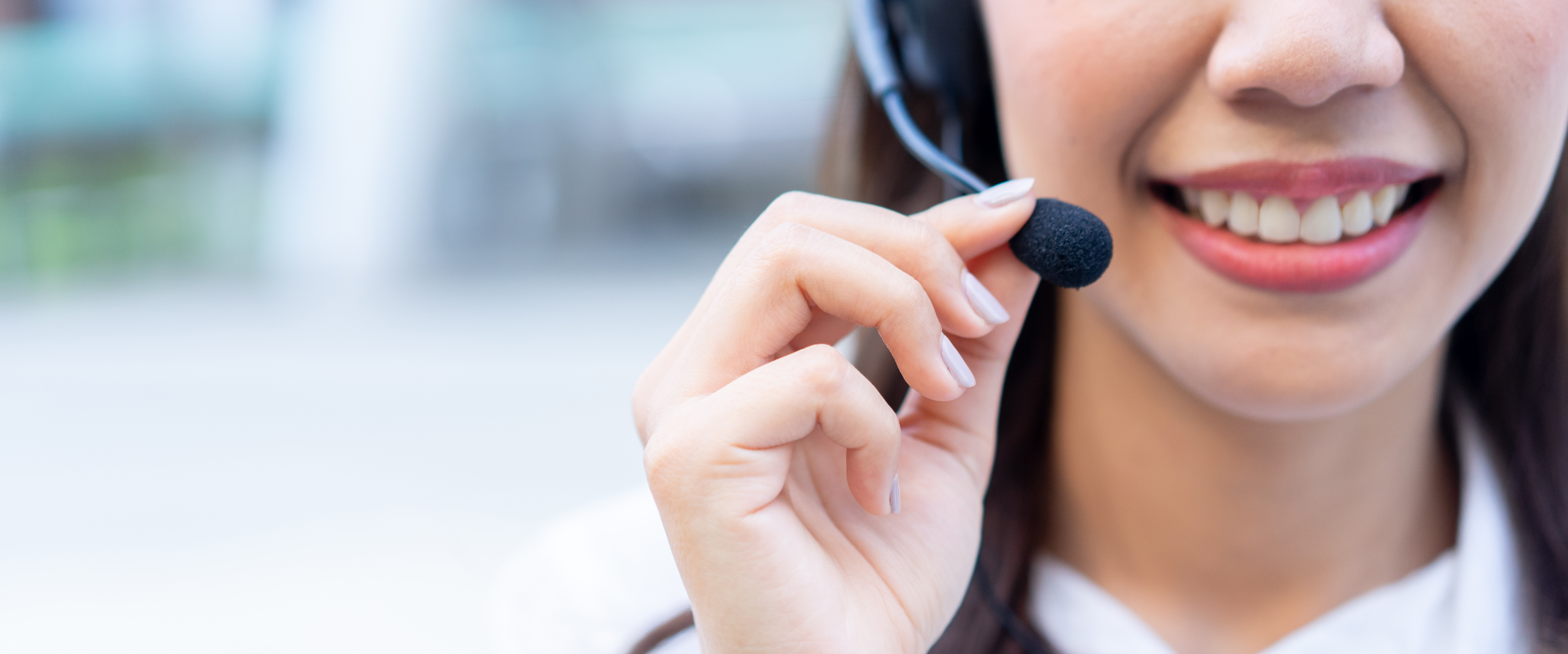 Close up photo showing the lower half of a woman's face. She is smiling and wearing a telephone headset. She is holding the microphone part with her right hand