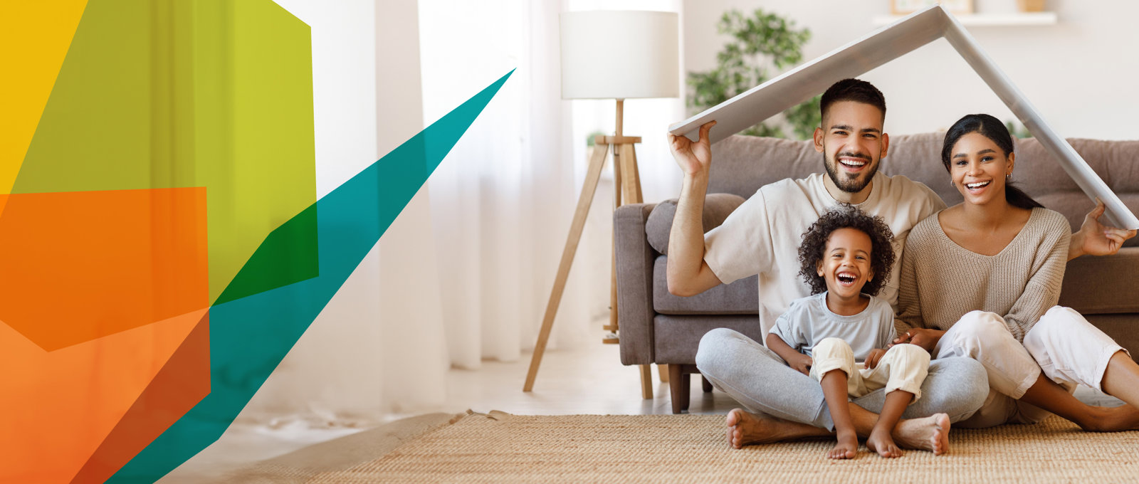 Photo of young mum, dad and child sitting in their living room. They are smiling and holding a folded board over their heads to look like a roof.