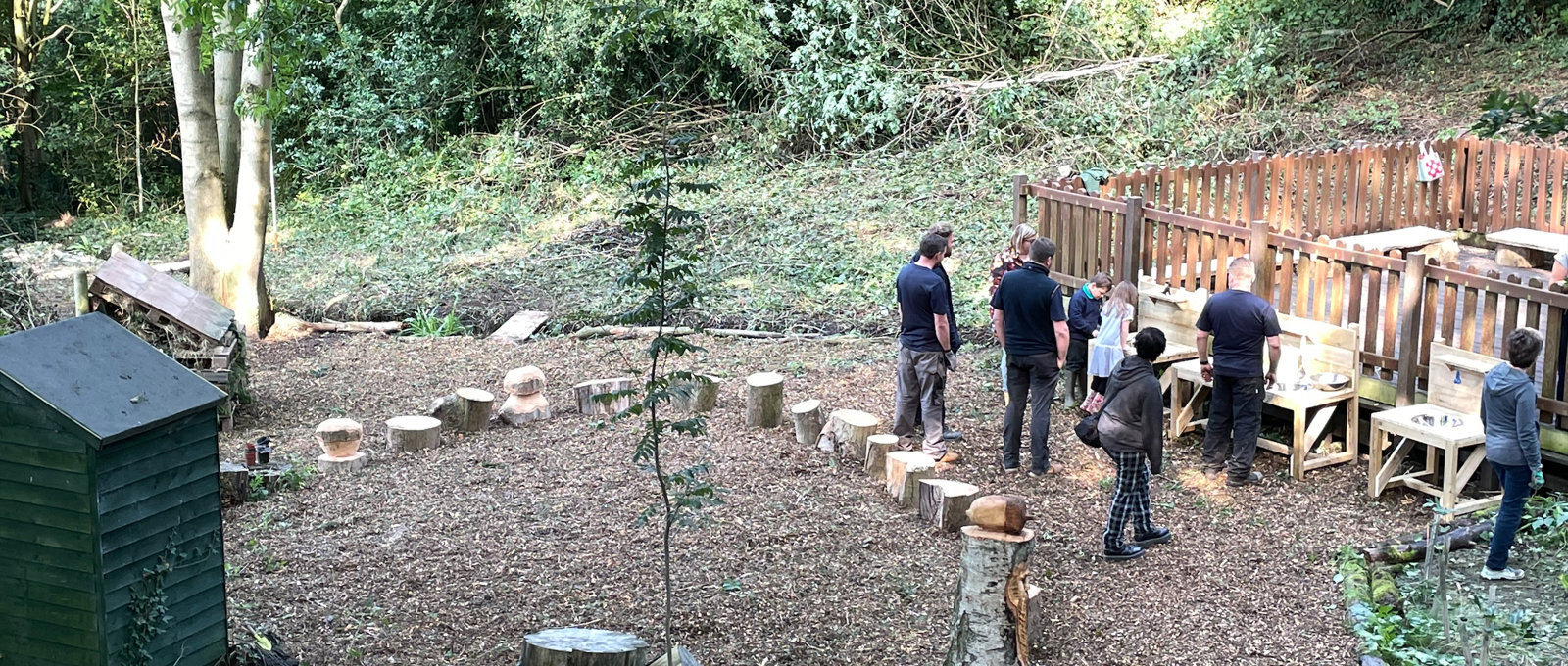 Photo of various team members from Futures working outdoors in a small woodland area at Lons infant School. There are various new wooden constructions in the photo.
