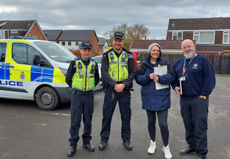 Photo of two Futures team members with two police officers, standing near some Futures homes with a police car in the background