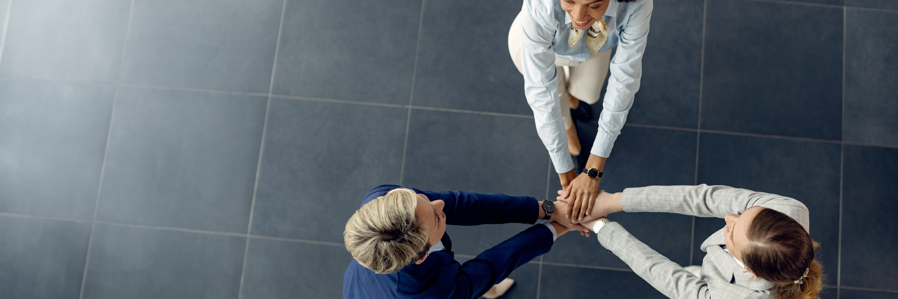 Overhead photo of three women, dressed in 'office wear'', standing in a circle clasping their hands together in the centre