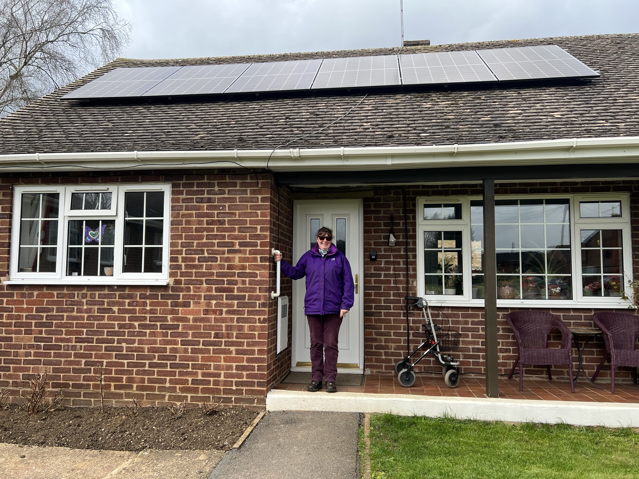 Photo of a Futures' customer called Lucille, standing on the front step outside her bungalow. She's wearing a purple coat and purple trousers, has dark hair tied up and sunglasses. Her bungalow has solar panels on the roof.