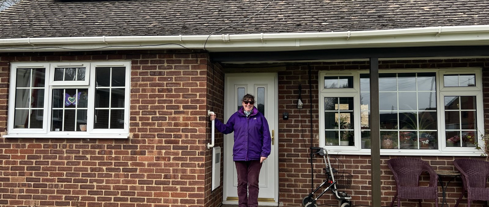 Photo of a Futures' customer called Lucille, standing on the front step outside her bungalow. She's wearing a purple coat and purple trousers, has dark hair tied up and sunglasses. Her bungalow has solar panels on the roof.