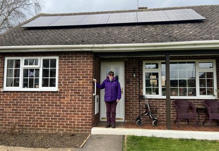 Photo of a Futures' customer called Lucille, standing on the front step outside her bungalow. She's wearing a purple coat and purple trousers, has dark hair tied up and sunglasses. Her bungalow has solar panels on the roof.
