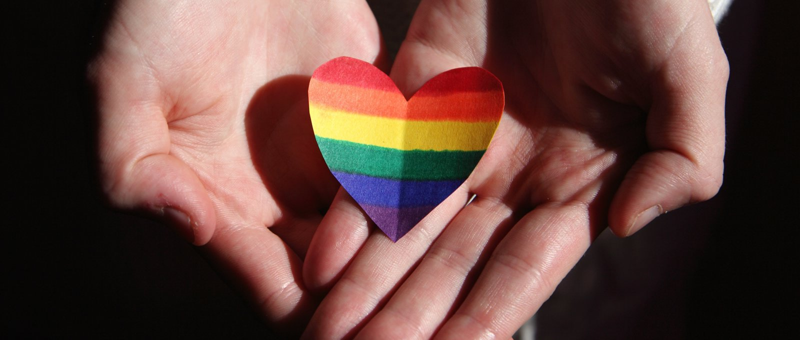 Close up photo of someone holding a cutout heart shape in rainbow colours in their outstretched, cupped hands