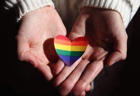 Close up photo of someone holding a cutout heart shape in rainbow colours in their outstretched, cupped hands