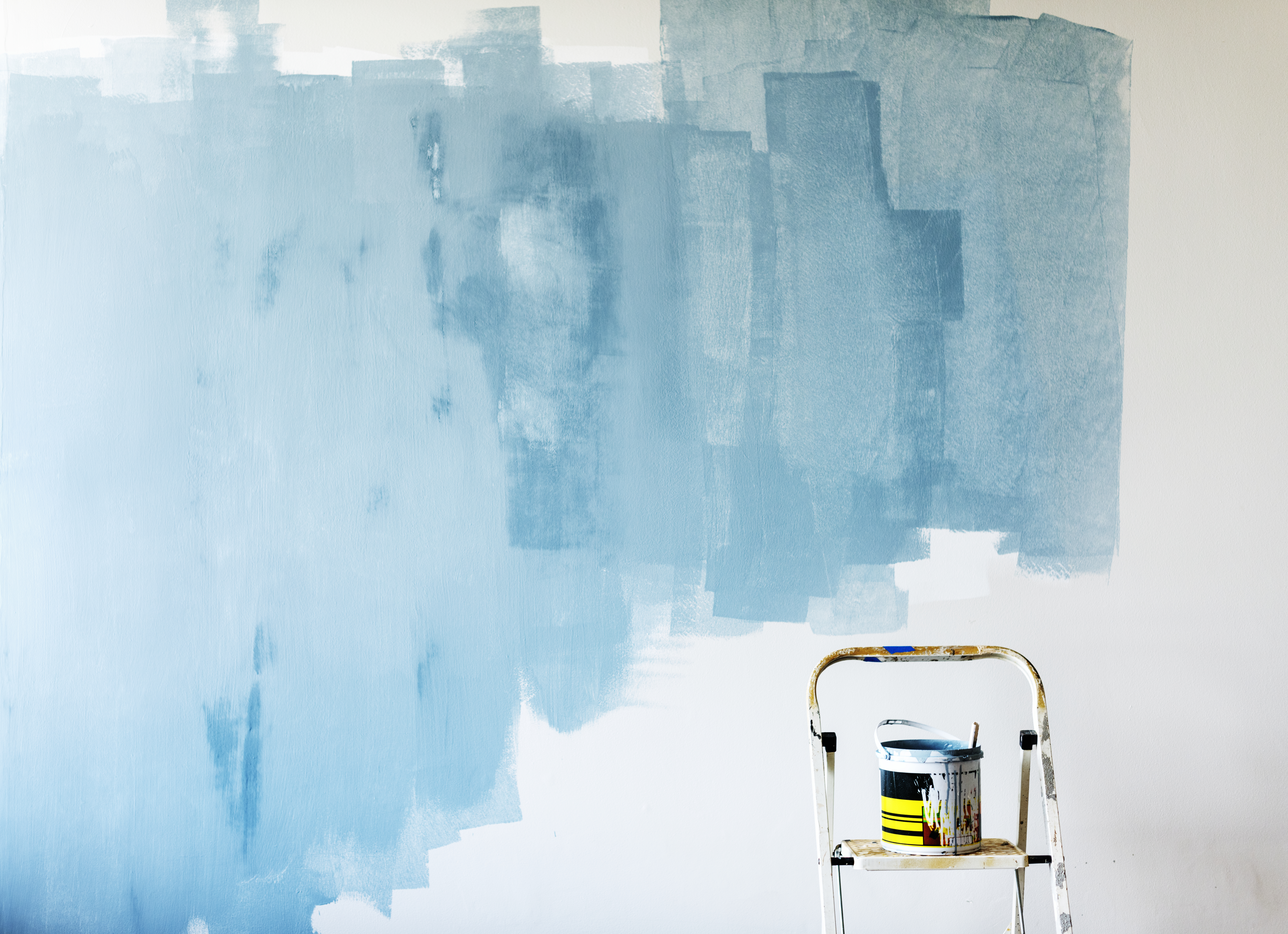 Photo of a white wall inside a house that someone has started to paint light blue. There is a stepladder in the bottom of the frame that has a pot of paint on it. 