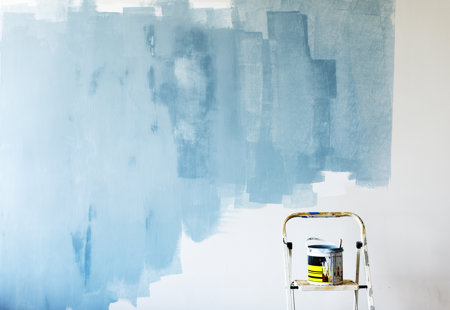 Photo of a white wall inside a house that someone has started to paint light blue. There is a stepladder in the bottom of the frame that has a pot of paint on it. 