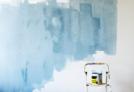 Photo of a white wall inside a house that someone has started to paint light blue. There is a stepladder in the bottom of the frame that has a pot of paint on it.