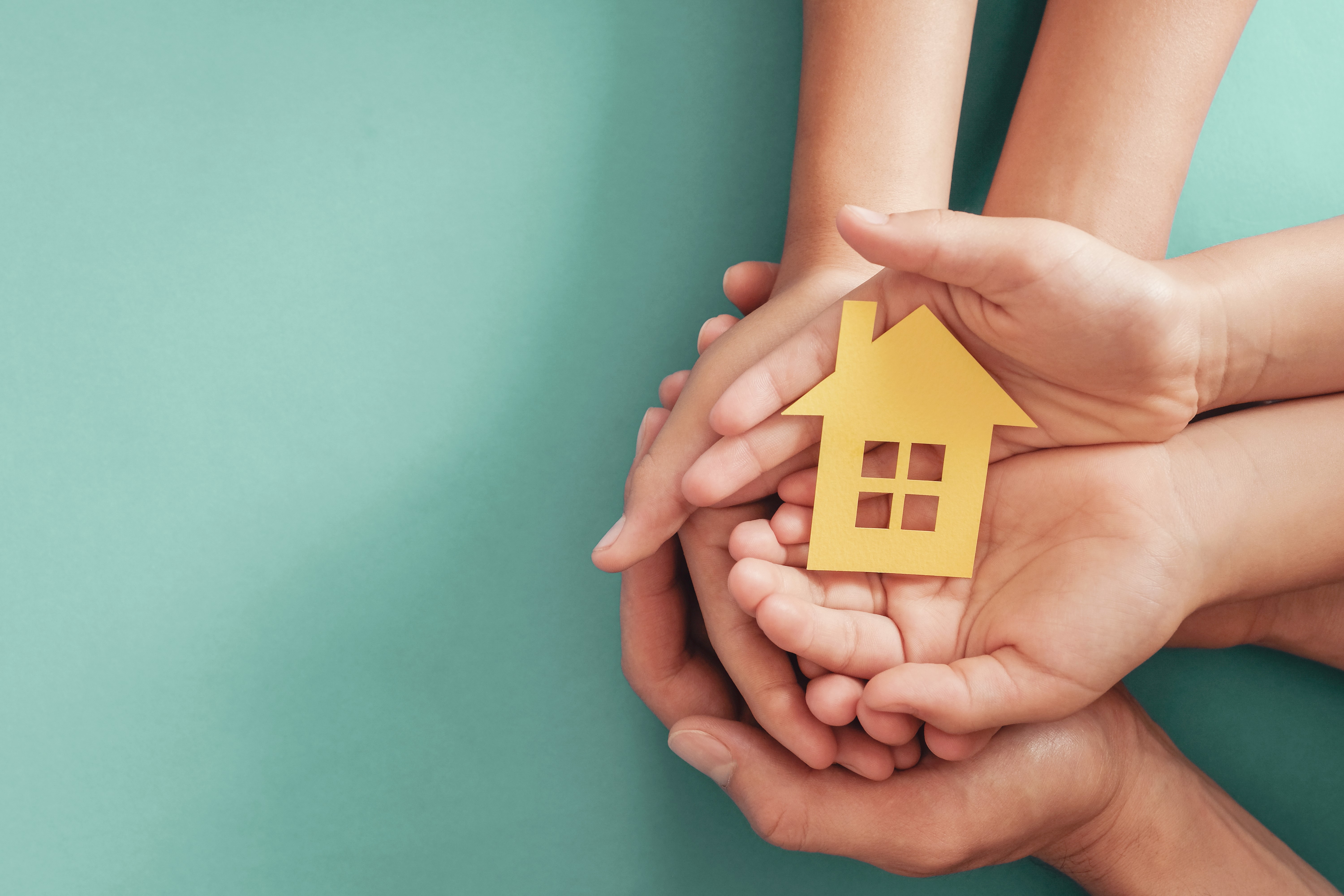 Photograph of several hands coming together to create a 'cup' shape and a small wooden cut-out of a house is being cradled in the hands