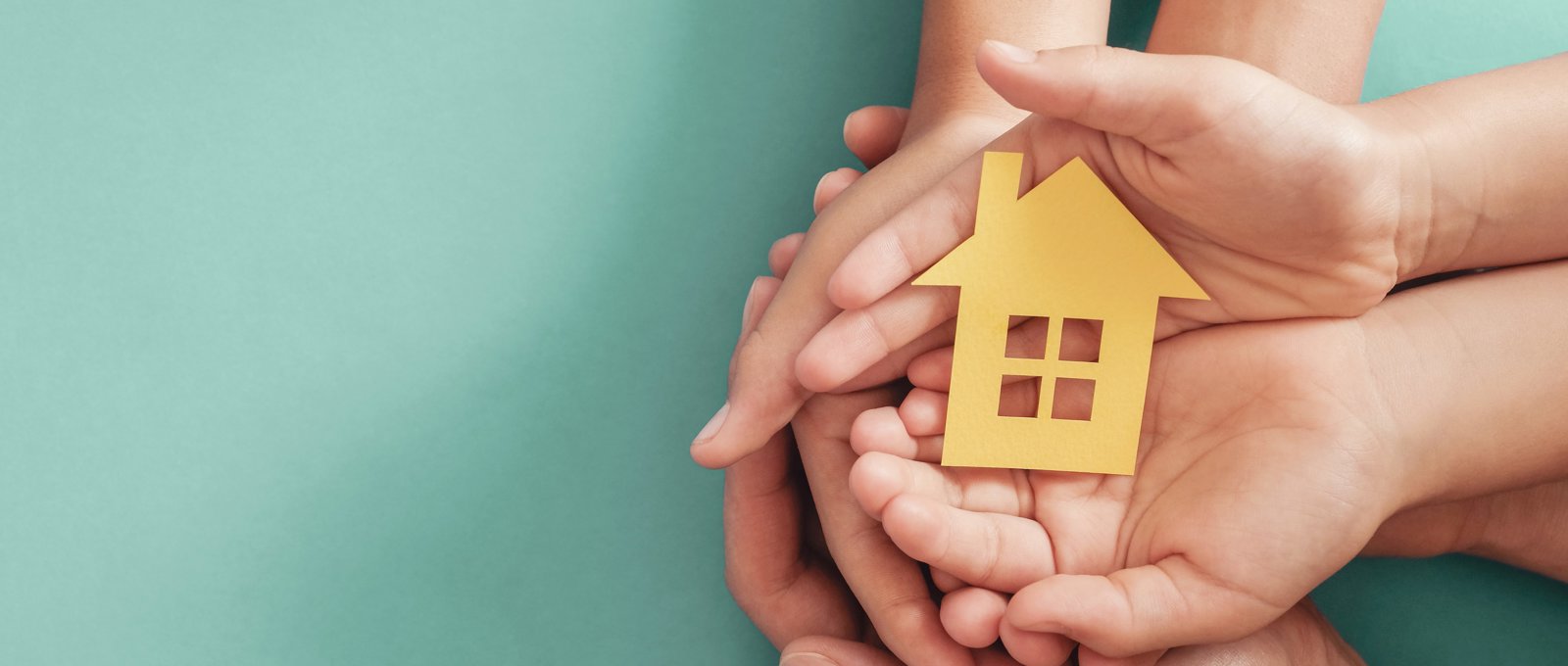 Photograph of several hands coming together to create a 'cup' shape and a small wooden cut-out of a house is being cradled in the hands