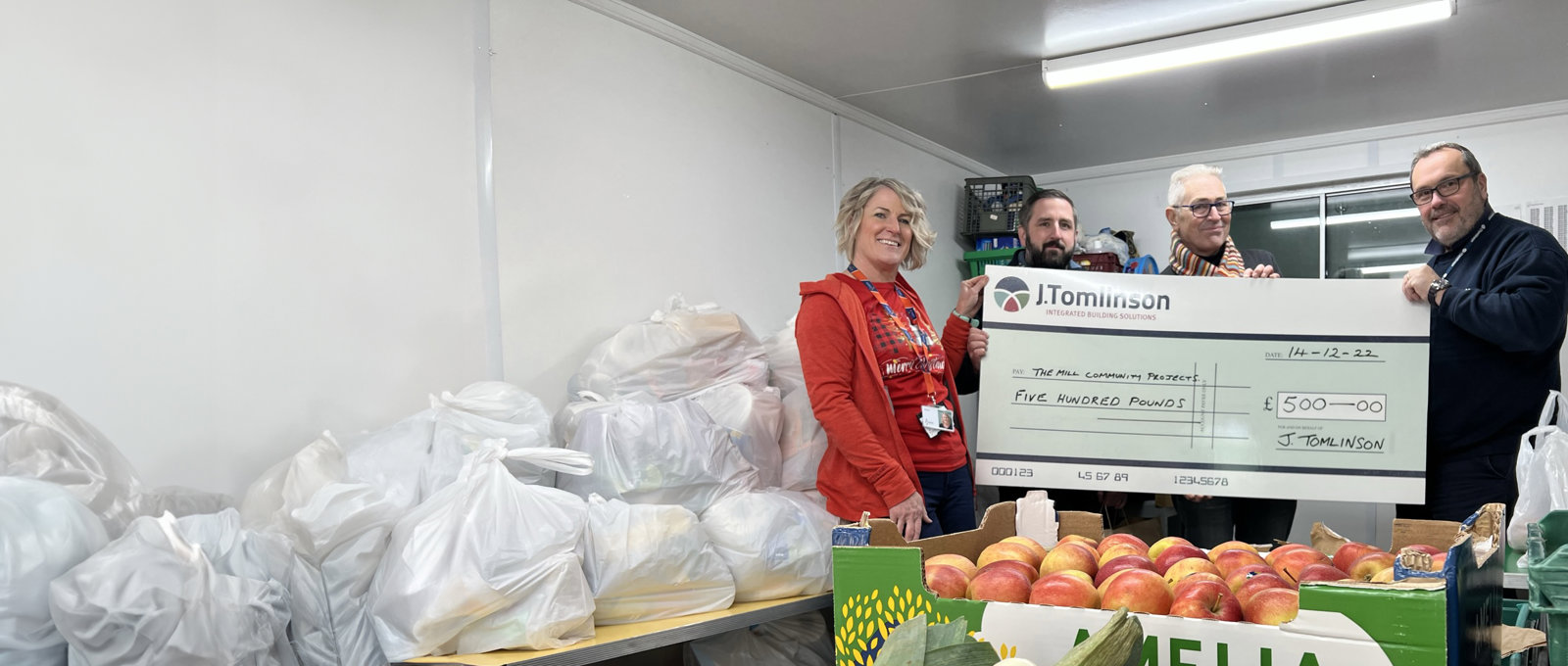 A photograph of four people holding a giant printed bank cheque and smiling to the camera. There are piles of fruit in the foreground and what look like food parcels behind them