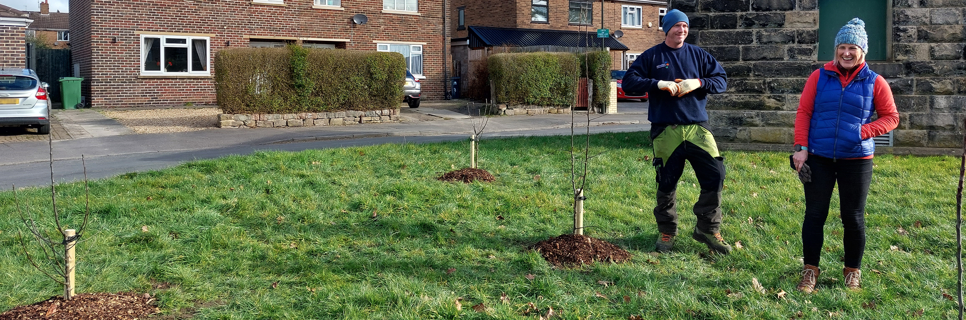 Photo of a man and a woman wearing winter clothes standing by some small, newly planted trees on a grassed area on a residential street